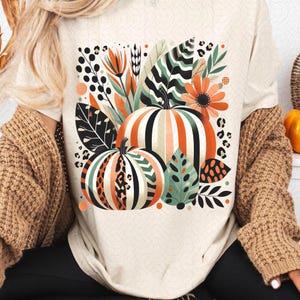 Peut inclure: Un t-shirt blanc avec un motif graphique de citrouilles, de feuilles et de fleurs dans des tons d'orange, de vert et de noir.