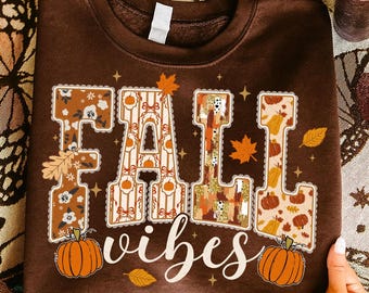 May include: A brown sweatshirt with the words "FALL vibes" in a decorative font. The letters are filled with fall-themed patterns, including pumpkins, leaves, and floral designs. Two orange pumpkins are at the bottom of the sweatshirt.
