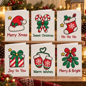 Op de afbeelding: Zes witte wenskaarten met geborduurde kersticonen: een kerstmuts met Merry Xmas, zuurstokken met Sweet Christmas, een kerstsok met Ho Ho Ho, een cadeau met Joy to You, wanten met Warm Wishes en maretak met Merry & Bright.