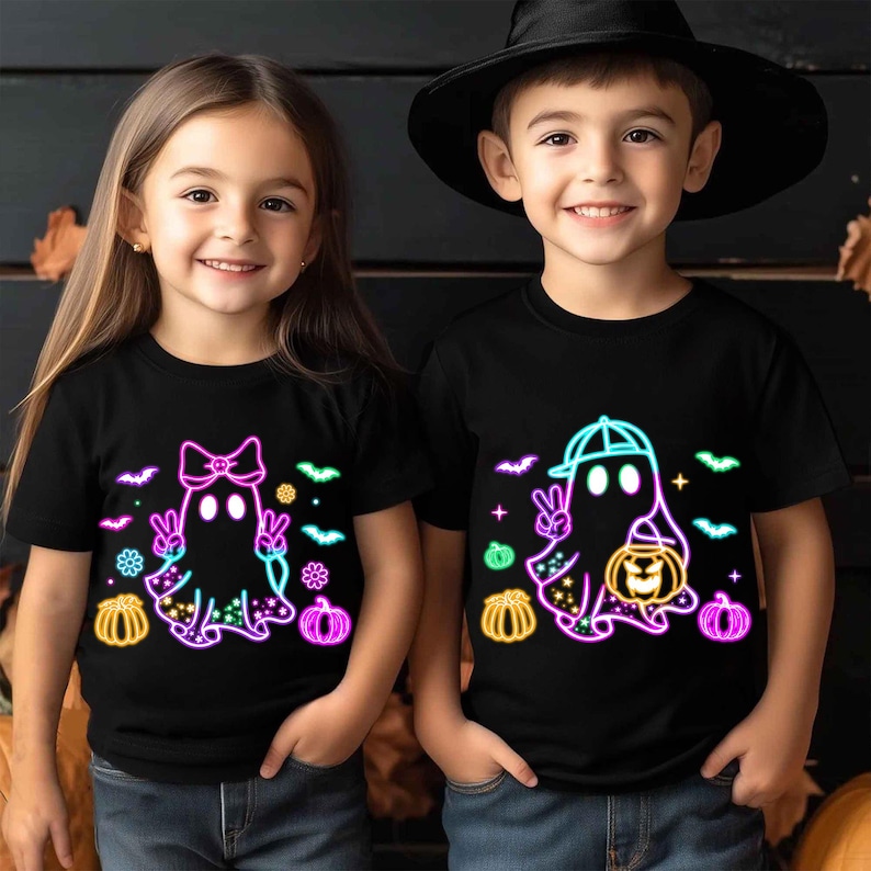 K&ouml;nnte beinhalten: Zwei Kinder tragen schwarze T-Shirts mit leuchtenden Geistermotiven. Der Geist des M&auml;dchens hat eine rosa Schleife und der Geist des Jungen eine blaue Baseballkappe. Beide Geister halten Jack-o'-Lantern.