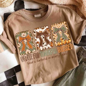 May include: A tan t-shirt featuring three decorative squares with bows, pumpkins, and floral designs. The words "Grateful, Thankful, Blessed" are printed across the front, along with the phrase "God is faithful in every season."