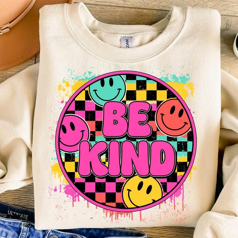 以下が含まれることがあります： スマイリーフェイスとチェック柄に囲まれたピンクの文字で「Be Kind」と書かれたカラフルなグラフィックデザインが施されたベージュのスウェットシャツ。
