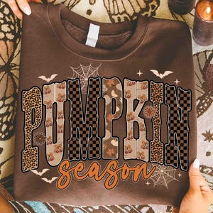 Puede incluir: Una sudadera marrón con las palabras "PUMPKIN season" en un diseño con temática otoñal. Las letras presentan estampado de leopardo, patrones de tablero de ajedrez y hojas de otoño. Murciélagos y telarañas también están incluidos.