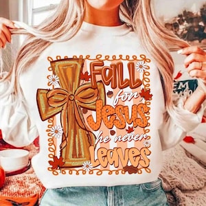 May include: White long-sleeve sweatshirt with a fall-themed graphic. The design features a cross with a bow, the words "Fall for Jesus," and "he never leaves." The design includes fall leaves and daisy accents.