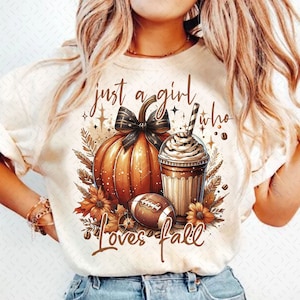 Puede incluir: Camiseta color crema con un diseño gráfico de temática otoñal. El diseño presenta una calabaza con un lazo negro, una bebida de café y un balón de fútbol. El texto dice "just a girl who loves fall."