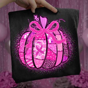 May include: Black t-shirt featuring a pink pumpkin design with a large bow and a pink ribbon. The pumpkin is composed of various pink patterns, including floral, plaid, and leopard print. The design is surrounded by a glittery border.