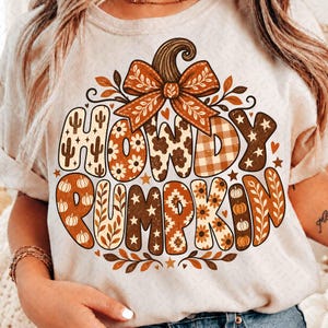 以下が含まれることがあります： クリーム色のTシャツで、リボンをあしらった大きなカボチャのグラフィックが特徴です。サボテン、花、牛柄、チェック、星などの様々な模様で埋め尽くされた大きな文字で「HOWDY PUMPKIN」と書かれており、小さなカボチャがアクセントになっています。