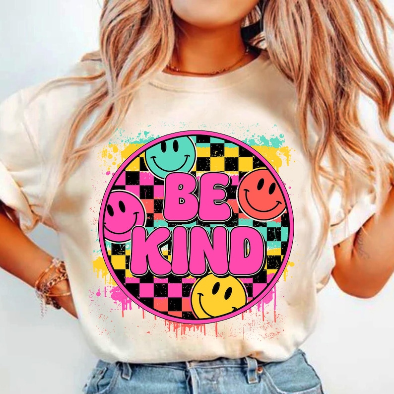 以下が含まれることがあります： スマイリーフェイスと市松模様に囲まれた「Be Kind」の文字が描かれたカラフルなグラフィックデザインの白いTシャツです。