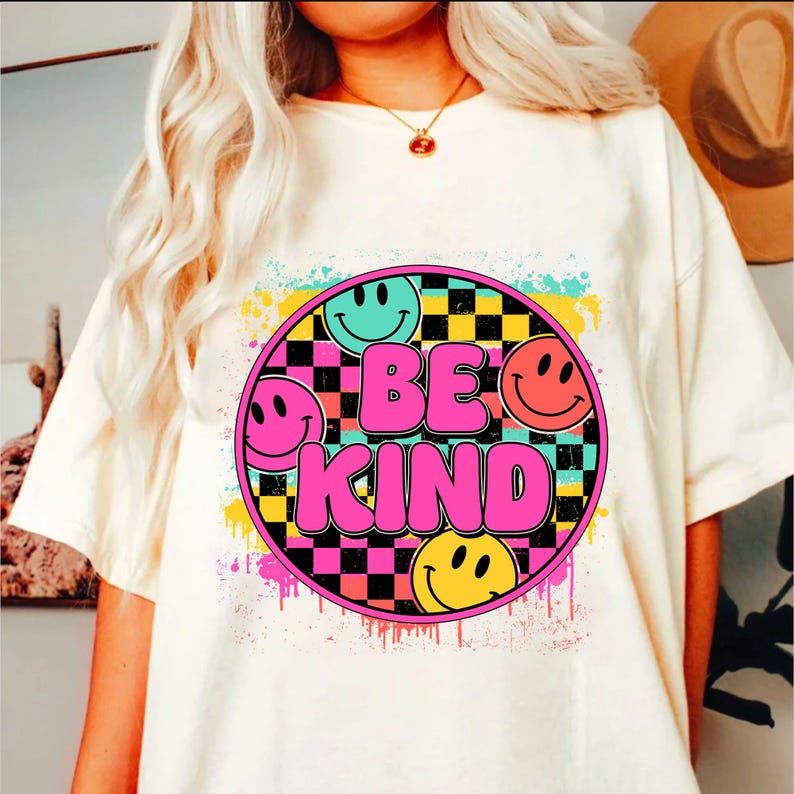 以下が含まれることがあります： 白地にカラフルなグラフィックデザインが施されたTシャツ。チェック柄の円の中にピンクの文字で「Be Kind」と書いてあり、4つのスマイリーフェイスが描かれている。