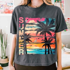 May include: Dark gray t-shirt with a colorful graphic of a sunset, palm trees, and seagulls. The word "SUMMER" is vertically printed on the left side of the design in a gradient of colors. The shirt has short sleeves and a crew neck.