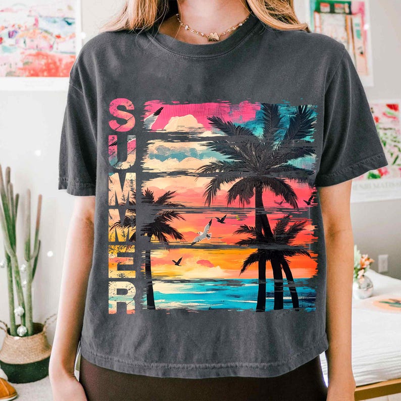 May include: Dark gray t-shirt with a colorful graphic of a sunset, palm trees, and seagulls. The word "SUMMER" is vertically printed on the left side of the design in a gradient of colors. The shirt has short sleeves and a crew neck.