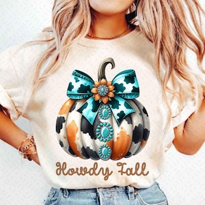 May include: A white t-shirt with a cow print pumpkin with a turquoise and black bow and turquoise rhinestones. The text "Howdy Fall" is written in brown glitter below the pumpkin.
