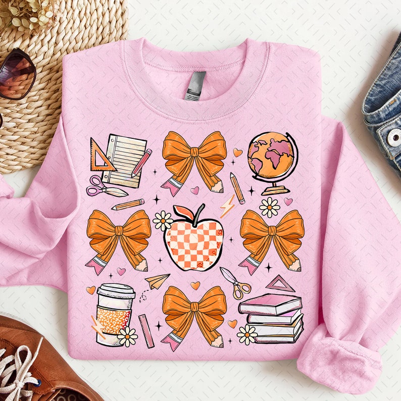 May include: Pink sweatshirt with a colorful design featuring a checkered apple, bows, pencils, a globe, a coffee cup, and other school supplies.