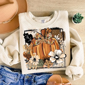 May include: Cream-colored sweatshirt featuring a large orange pumpkin design with a decorative bow and floral accents. The pumpkin is set within a black rectangular frame, with additional flowers and leaves surrounding it. The sweatshirt is paired with denim shorts and sandals.