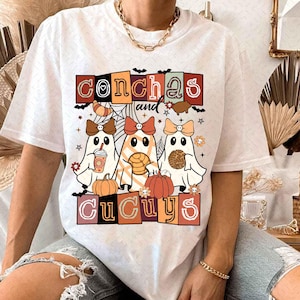 May include: White t-shirt with a graphic design featuring three ghosts holding a pumpkin, a coffee cup, and a pastry. The text "conchas and cucuys" is written in a colorful block font.