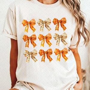 May include: A white t-shirt with a pattern of nine orange and yellow bows. The bows are in a variety of styles, including plaid, floral, and solid.