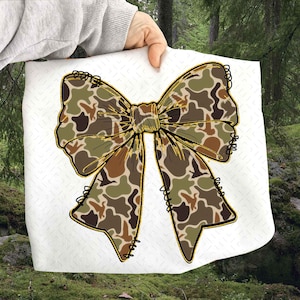 May include: A large, decorative bow with a camouflage pattern in shades of green, brown, and tan. The bow has a gold outline and black stitching details. The bow is displayed on a white background.