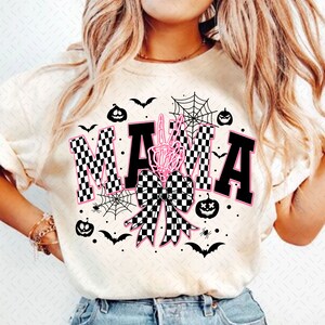 May include: White t-shirt with a black and white checkered "MAMA" graphic with a pink skeleton hand and a black and white checkered bow. The graphic is surrounded by black bats, spiderwebs, and pumpkins.