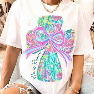 Könnte beinhalten: Weißes T-Shirt mit einem bunten Blumenkreuz-Design und dem Text "He is Risen".