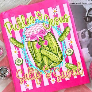 May include: Pink blanket with white vertical stripes and the words "Pickles & Jesus" and "Salty & Saved" in a whimsical font. A jar of pickles with a pink bow is the central image, surrounded by pickle illustrations and daisies.