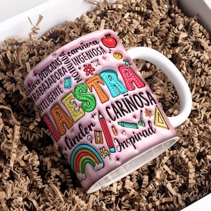 May include: Pink ceramic mug with a colorful design that says "Maestra" and "Linda + Cari&ntilde;osa + Inspirad". The mug has a white handle and is surrounded by brown packing paper.