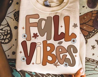May include: Cream-colored sweatshirt with the words "FALL VIBES" in brown, tan, and cream lettering. The design includes fall leaves, daisies, and stars. The sweatshirt is folded and held by hands.