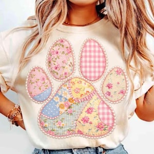 May include: Cream-colored t-shirt featuring a patchwork paw print design. The paw print is composed of floral and gingham patterns in pink, blue, and yellow, outlined with a pearl-like trim. A charming and unique design.