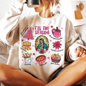 May include: A white sweatshirt with a festive design featuring the text "Tis the Season" and illustrations of traditional Mexican Christmas foods and drinks, including tamales, churros, champurrado, menudo, pan dulce, and a Christmas tree.
