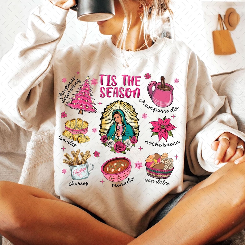May include: A white sweatshirt with a festive design featuring the text "Tis the Season" and illustrations of traditional Mexican Christmas foods and drinks, including tamales, churros, champurrado, menudo, pan dulce, and a Christmas tree.