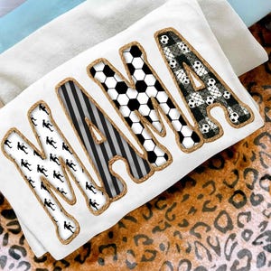 Op de afbeelding: Een wit kussen met het woord "MAMA" in grote letters. Elke letter heeft een ander ontwerp: voetballers, zwart-witte strepen, voetballen en een zwart-wit kanten patroon. De letters zijn omlijnd met een touwachtige rand.