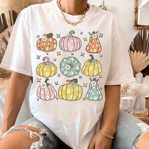 May include: White t-shirt with a colorful pattern of pumpkins with flowers. The pumpkins are in various shades of orange, yellow, green, and blue.