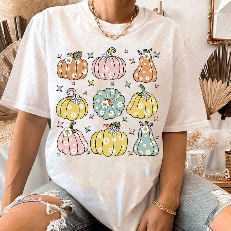 May include: White t-shirt with a colorful pattern of pumpkins with flowers. The pumpkins are in various shades of orange, yellow, green, and blue.