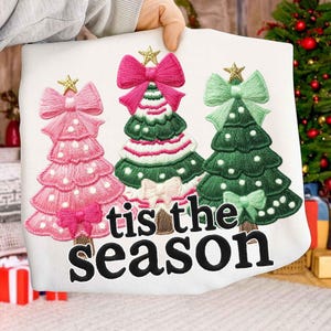 Puede incluir: Sudadera blanca con temática navideña con árboles de Navidad bordados en rosa, verde y blanco, cada uno coronado con una estrella dorada y un lazo grande. La frase "tis the season" está bordada en negro debajo.