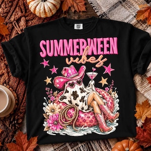 Puede incluir: Una camiseta negra con un estampado gráfico que incluye el texto "SUMMERWEEN vibes" en rosa y naranja. El diseño muestra un fantasma con sombrero de vaquera rosa y gafas de sol en forma de corazón, sentado sobre un flotador rosa con flores.