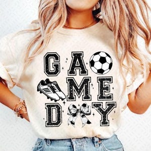 May include: White t-shirt with black glitter text that reads "GAME DAY" with a soccer ball, soccer cleats, and a bow with a soccer ball pattern.