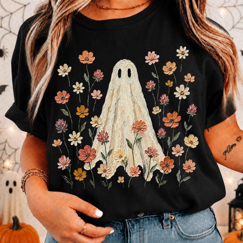 May include: A black t-shirt featuring a central graphic of a cream-colored ghost with a textured, impasto-style appearance. The ghost is surrounded by a variety of small, colorful flowers in shades of pink, orange, yellow, and white.