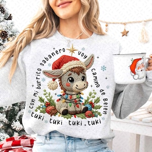 May include: A light gray sweatshirt featuring a cartoon donkey wearing a Santa hat and Christmas lights. The donkey is surrounded by festive decorations and text. A white mug with a Santa Claus illustration is held in the hand.