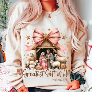 May include: A cream-colored sweatshirt with a pink bow and a nativity scene graphic. The graphic includes a manger, Mary, Joseph, baby Jesus, and sheep. The text "Greatest Gift of All" and "Matthew 1:21" is printed below the graphic.