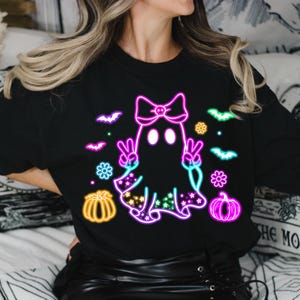 K&ouml;nnte beinhalten: Schwarzes T-Shirt mit einem neonrosa, blauen und gr&uuml;nen Geist, der eine rosa Schleife mit einem Totenkopfdesign tr&auml;gt. Der Geist h&auml;lt zwei Peace-Zeichen hoch. Auf dem Shirt befinden sich auch neonrosa, blaue und gr&uuml;ne K&uuml;rbisse und Flederm&auml;use.