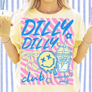 May include: Yellow t-shirt with a pink and white wavy design and the words "DILLY DILLY CLUB members only" in blue. The shirt features a smiley face, a bow, and a drink graphic.