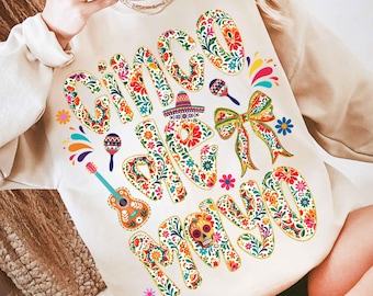 May include: A cream-colored sweatshirt with the words "Cinco de Mayo" in a colorful floral design. The design includes a sombrero, maracas, a guitar, and a decorative bow, all in vibrant colors.
