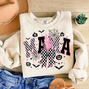 May include: A cream colored sweatshirt with a black and white checkered bow and the word "MAMA" in black letters with pink outlines. The design also includes a skeleton hand, bats, spiders, and pumpkins.