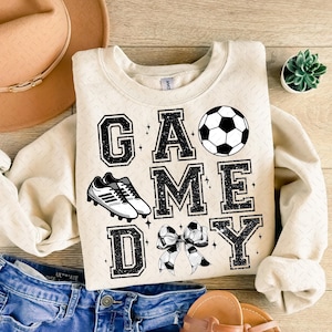 May include: A beige sweatshirt with the text "GAME DAY" in black with a soccer ball, soccer cleats, and a bow.