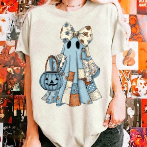 Op de afbeelding: Illustratie van een lichtblauw spook met een patchwork-ontwerp, met een strik en een blauwe pompoenmand. Het spook heeft zwarte ogen en is versierd met bloemmotieven, perfect voor Halloween.