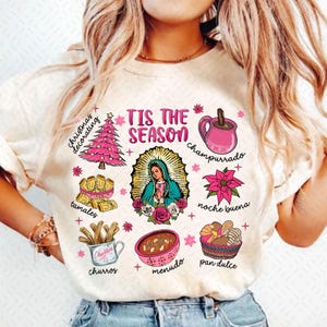 May include: A white t-shirt with a pink and white design featuring the text "Tis the Season" and various Mexican Christmas traditions, including tamales, churros, champurrado, and pan dulce.