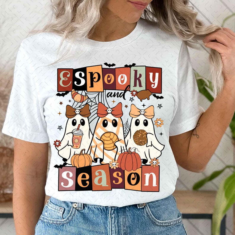 Pu&ograve; includere: Maglietta bianca con una grafica di tre fantasmi che tengono oggetti autunnali. Il testo "E Spooky and Season" &egrave; in un font retr&ograve;.