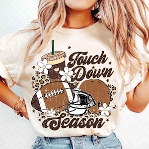 May include: White t-shirt with a brown football, helmet, coffee cup, and flowers. The text "Touch Down Season" is printed on the shirt.