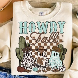 May include: A cream coloured sweatshirt with the text "Howdy Fall" in brown and blue lettering. Three ghosts wearing cowboy hats are standing in front of a chequered background with cacti on either side. The ghosts are wearing cow print, brown and white, and blue and white patterns.