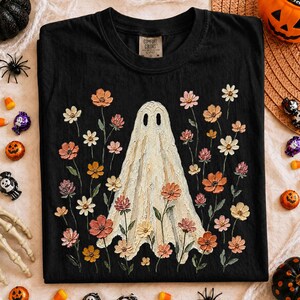 May include: A black short-sleeve t-shirt featuring a central graphic of a cream-colored ghost surrounded by various colorful flowers in shades of pink, orange, and white. The shirt has a crew neckline and a Comfort Colors brand label at the neck.
