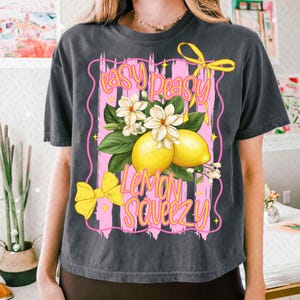 Op de afbeelding: Een zwart getailleerd t-shirt met een roze en wit grafisch ontwerp met citroenen, bloemen en de tekst "easy peasy lemon squeezy".